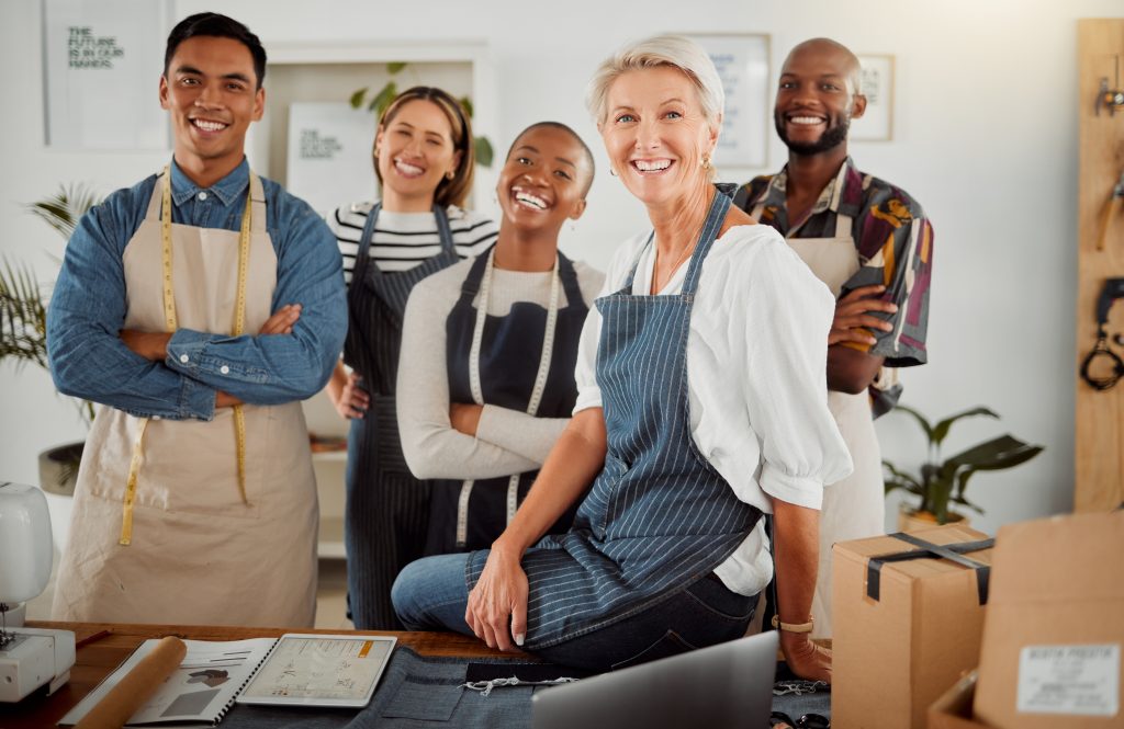 Group of five cheerful diverse clothing designers standing with their arms crossed in a shop at wor.